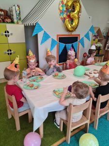Dzieci siedzą przy urodzinowym stoliku i częstują się urodzinowymi babeczkami. W tle widać urodzinową dekorację.