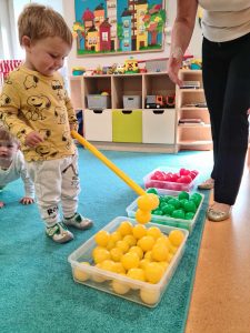 Chłopiec stoi na turkusowym dywanie. Trzyma w rączce żółtą tubę. Przed nim na dywanie leżą trzy kwadratowe pojemniki wypełnione kolorowymi piłeczkami.