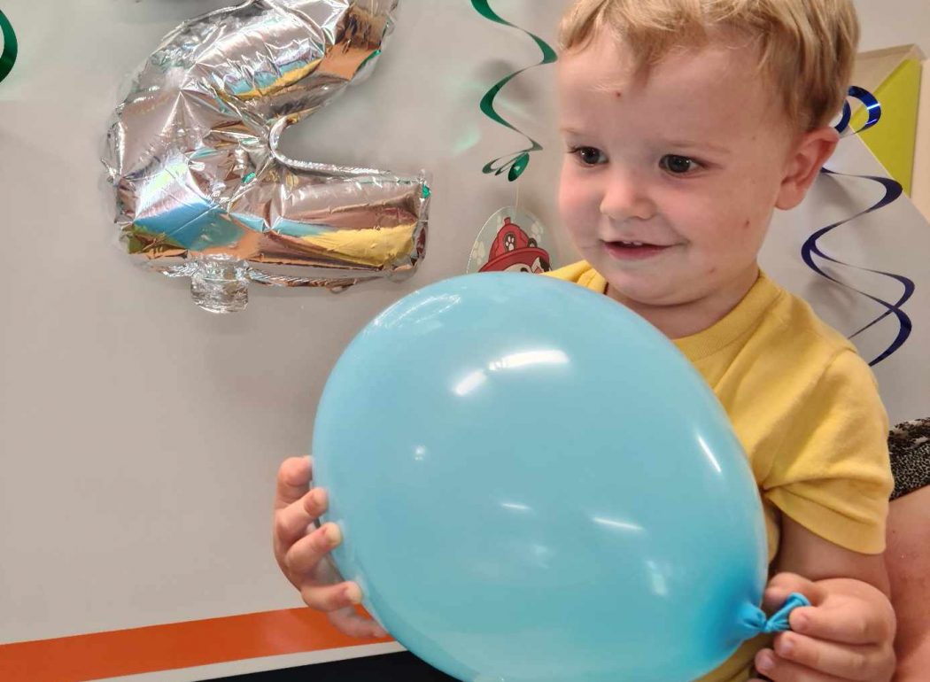 Uśmiechnięty chłopczyk trzyma w rączkach niebieski balon. W tle urodzinowe dekoracje i srebrny balon w kształcie cyferki dwa.