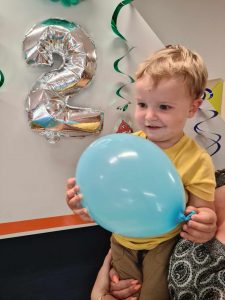 Uśmiechnięty chłopczyk trzyma w rączkach niebieski balon. W tle urodzinowe dekoracje i srebrny balon w kształcie cyferki dwa.