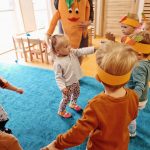 Dzieci tańczą na turkusowym dywanie z dużą, pomarańczową maskotką w kształcie marchewki.