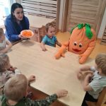 Dzieci razem z panią opiekunką i dużą pomarańczową maskotką w kształcie marchewki siedzą na krzesełkach przy stole. Dzieci patrzą na panią opiekunkę która trzyma w rękach talerz z poszatkowaną marchewką.