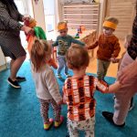 Dzieci tańczą w kółeczku razem z paniami opiekunkami na turkusowym dywanie.