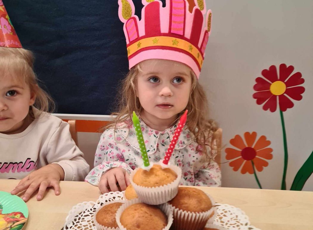 Dziewczynka siedzi przy stole, na głowie ma różową, urodzinową koronę. Przed nią na stole leży talerz z babeczkami.