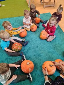 Dzieci siedzą na turkusowym dywanie, trzymają w dłoniach pomarańczowe dynie.