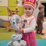 Dziewczynka stoi na turkusowym dywanie. Na głowie ma różową, urodzinową koronę a w dłoniach trzyma balon w kształcie cyfry dwa.