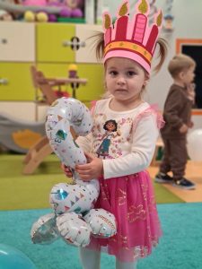 Dziewczynka stoi na turkusowym dywanie. Na głowie ma różową, urodzinową koronę a w dłoniach trzyma balon w kształcie cyfry dwa.