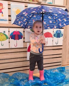 Dziewczynka stoi na niebieskiej foli rozłożonej na podłodze, ma na nogach różowe kalosze i trzyma w rączkach granatowy parasol. W tle widać przyklejone na ścianie obrazki z kolorowymi parasolami.
