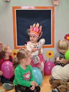 Dziewczynka siedzi na krzesełku, na głowie ma różową urodzinową koronę a w dłoniach trzyma balon w kształcie cyfry dwa. Wokół niej siedzą dzieci, trzymają w dłoniach kolorowe baloniki.
