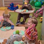 Dzieci siedzą na krzesełkach przy stoliku i jedzą babeczki. Na stole leży duży talerz z babeczkami, małe, kolorowe, papierowe talerzyki i kolorowe plastikowe kubeczki.