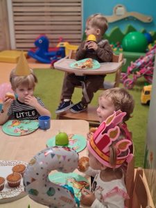 Dzieci siedzą na krzesełkach przy stoliku i jedzą babeczki. Na stole leży duży talerz z babeczkami, małe, kolorowe, papierowe talerzyki i kolorowe plastikowe kubeczki.