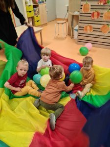 Dzieci siedzą na rozłożonej na podłodze kolorowej chuście animacyjnej i bawią się kolorowymi balonikami.
