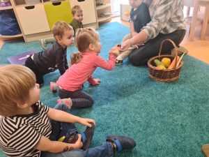 Dzieci siedzą na turkusowym dywanie. Naprzeciwko nich na dywanie siedzi pani opiekunka, obok niej leży wiklinowy koszyk z owocami i warzywami. Pani opiekunka pokazuje dzieciom co znajduje się w koszyku.