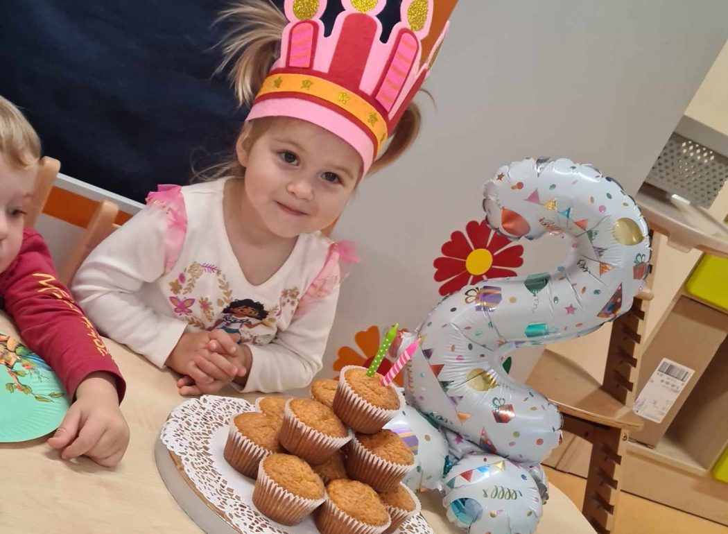 Dziewczynka siedzi na krzesełku przy stole, na głowie ma urodzinową koronę. Przed nią na stole leży talerz z babeczkami i urodzinowy balon w kształcie cyfry dwa.
