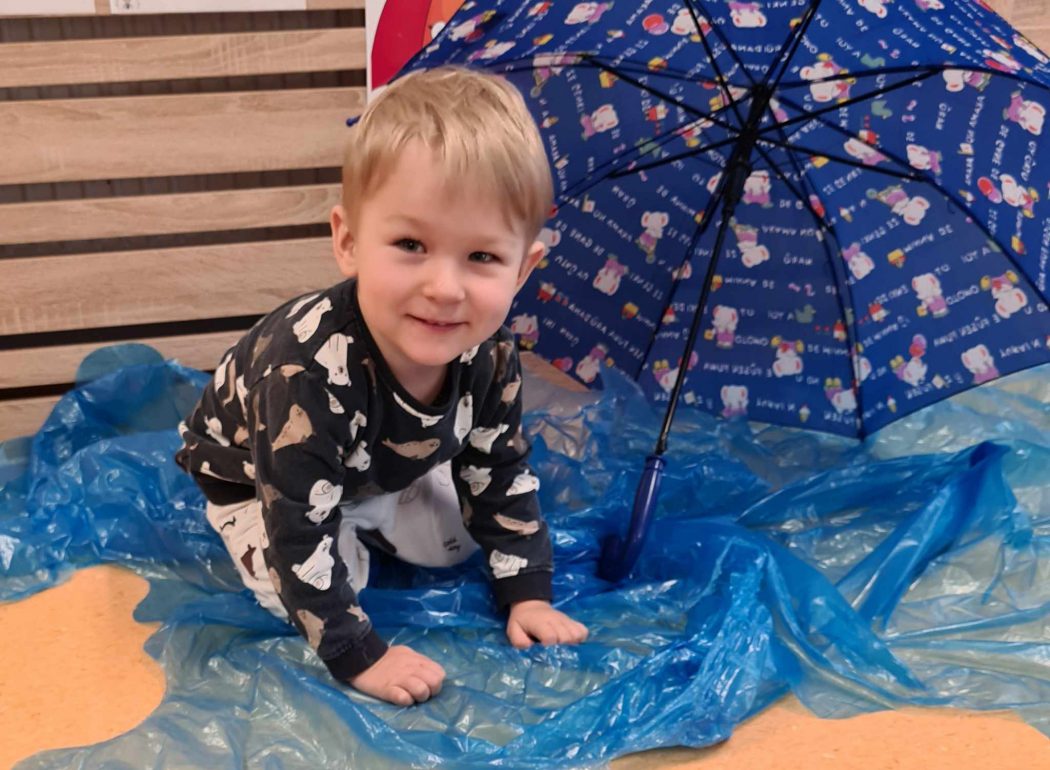 Chłopiec siedzi na niebieskiej foli rozłożonej na podłodze, obok niego leży rozłożony granatowy parasol.