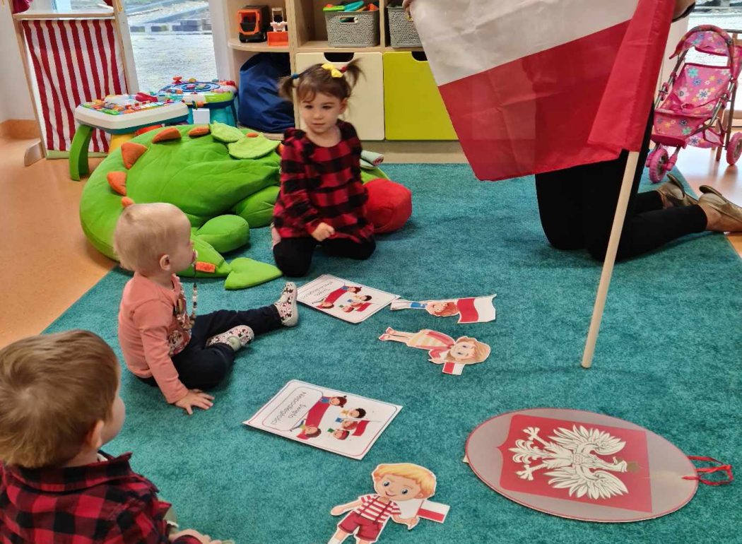 Dzieci siedzą na turkusowym dywanie i patrzą na flagę Polski, którą pokazuje im opiekunka. Na dywanie leży godło Polski i grafiki przedstawiające symbole narodowe.