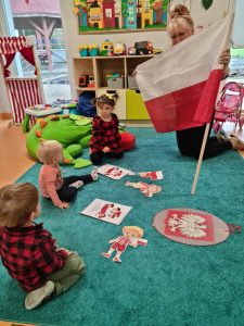 Dzieci siedzą na turkusowym dywanie i patrzą na flagę Polski, którą pokazuje im opiekunka. Na dywanie leży godło Polski i grafiki przedstawiające symbole narodowe.