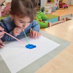 Dziewczynka siedzi na krzesełku przy stole, rozdmuchuje za pomocą kolorowej słomki niebieską farbę na kartce papieru.