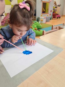 Dziewczynka siedzi na krzesełku przy stole, rozdmuchuje za pomocą kolorowej słomki niebieską farbę na kartce papieru.