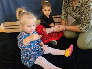 Dwie dziewczynki razem z opiekunką siedzą na granatowym materiale rozłożonym na podłodze. Dziewczynki losują karteczki z wróżbami z kolorowego pudełka.