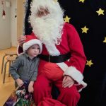 Mikołaj siedzi na krześle, obok niego stoi chłopiec, który ma na głowie mikołajową czapkę a w rączkach trzyma kolorową torbę z prezentem.