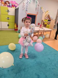 Dziewczynka stoi na turkusowym dywanie, trzyma w dłoniach balon w kształcie cyfry dwa. Na dywanie leżą kolorowe balony. W tle widać salę zabaw.