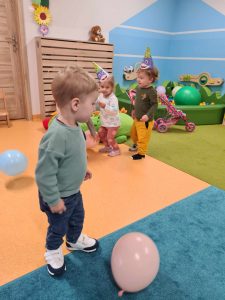 Dzieci tańczą na sali zabaw. Na podłodze leżą kolorowe baloniki.