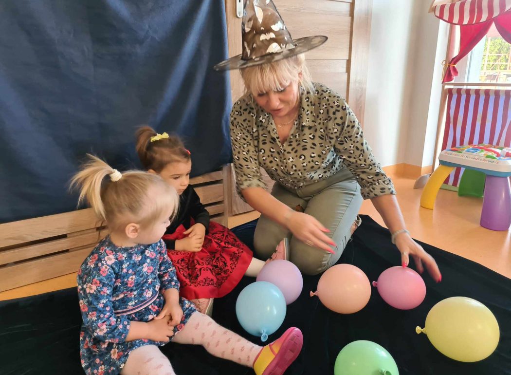 Dwie dziewczynki ubrane w sukienki, siedzą razem z opiekunką na granatowym materiale rozłożonym na podłodze. Opiekunka ma na głowie kapelusz. Na podłodze leżą kolorowe baloniki.