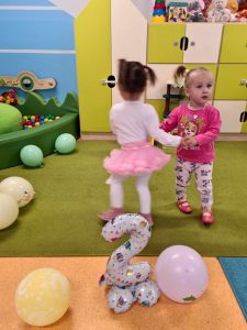 Dwie dziewczynki tańczą razem trzymając się za raczki. Na podłodze leżą kolorowe balony i balon w kształcie cyfry dwa. W tle widać salę zabaw.