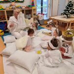 Ubrane na biało dzieci siedzą na białym kocu rozłożonym na podłodze, wokół nich leżą rozrzucone białe poduszki. Dzieci patrzą na opiekunkę która trzyma w dłoniach figurkę anioła. W tle widać salę zabaw.