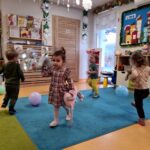Dzieci tańczą na turkusowym dywanie, między nimi na podłodze leżą kolorowe balony. W tle widać salę zabaw.