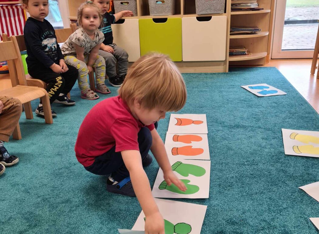 Chłopiec kuca na turkusowym dywanie i układa grafiki przedstawiające kolorowe czapki i rękawiczki. W tle widać dzieci siedzące na krzesełkach.
