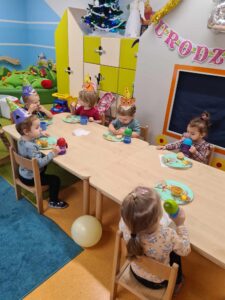 Dzieci siedzą przy stole, jedzą babeczki i piją wodę z kolorowych kubeczków. Część dzieci ma na głowach kolorowe, urodzinowe czapeczki. W tle widać salę zabaw.