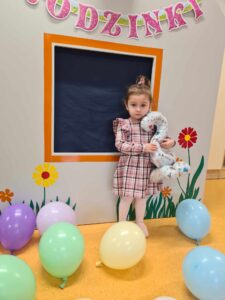 Dziewczynka ubrana w różową sukienkę w kratkę stoi i trzyma w dłoniach balon w kształcie cyfry dwa. Przed nią na podłodze leżą kolorowe balony. W tle widać szary domek ozdobiony kolorowymi kwiatkami.