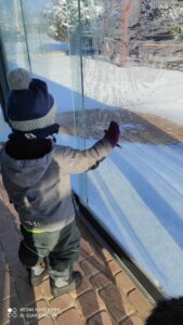 Chłopiec ubrany w czapkę, kurtkę i rękawiczki, stoi przy oknie i patrzy na zimowy krajobraz.