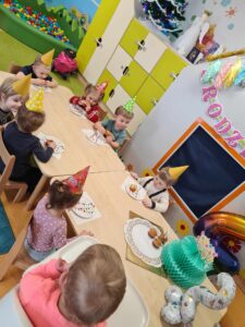 Dzieci siedzą na krzesełkach przy stole, na głowach mają kolorowe, urodzinowe czapeczki. Na stole leży talerz z babeczkami i urodzinowa dekoracja w kształcie tortu. Dzieci jedzą babeczki. W tle widać sale zabaw ozdobioną urodzinowymi dekoracjami.