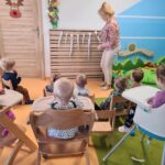 Na zdjęciu widać dzieci siedzące tyłem na krzesełkach. Dzieci patrzą na opiekunkę, która stoi naprzeciwko nich i pokazuje im zawieszone na ścianie sople wykonane z foli aluminiowej.