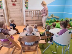 Na zdjęciu widać dzieci siedzące tyłem na krzesełkach. Dzieci patrzą na opiekunkę, która stoi naprzeciwko nich i pokazuje im zawieszone na ścianie sople wykonane z foli aluminiowej.