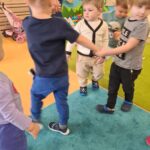 Dzieci tańczą w kole trzymając się za ręce. W tle widać salę zabaw.