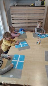 Dzieci siedzą przy stole i przyklejają białe papierowe śnieżynki na niebieską kartkę papieru.