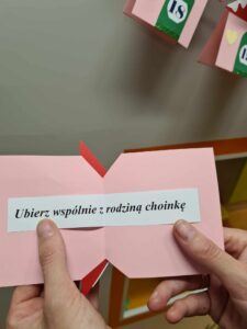 Na zdjęciu widać różowo-białą kartkę trzymaną w dłoniach. Na kartce napisane jest zadanie: " Ubierz wspólnie z rodziną choinkę".