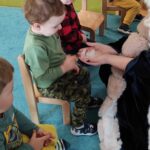 Dzieci siedzą na krzesełkach na turkusowym dywanie, oglądają kostki lodu, które trzyma w dłoniach opiekunka