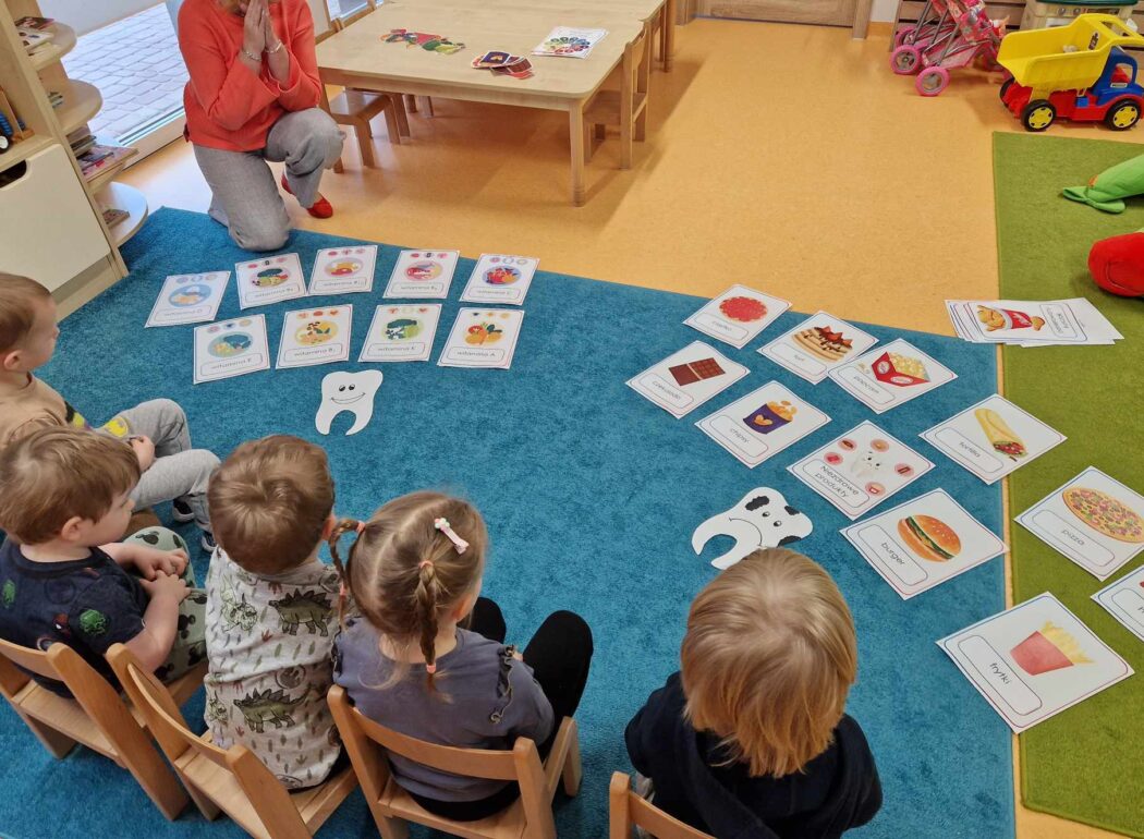 Dzieci siedzą na krzesełkach, patrzą na rozłożone na dywanie kolorowe grafiki przedstawiające zdrową i niezdrową żywność.