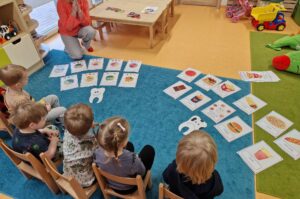 Dzieci siedzą na krzesełkach, patrzą na rozłożone na dywanie kolorowe grafiki przedstawiające zdrową i niezdrową żywność.
