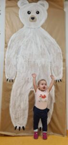 Dziewczynka stoi i trzyma ręce w górze. W tle widać dużego niedźwiedzia polarnego namalowanego farbami na szarym papierze.