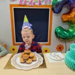 Chłopiec ubrany w urodzinową czapeczkę, siedzi przy stole i dmucha świeczkę na urodzinowym torcie zrobionym z babeczek. W tle widać szary domek ozdobiony urodzinowymi dekoracjami i duży kolorowy balon w kształcie cyfry trzy