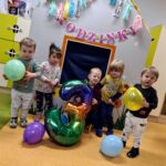 Dzieci stoją i pozują do zdjęcia z solenizantem, który siedzi na krzesełku i trzyma w dłoniach kolorowy, duży balon w kształcie cyfry trzy. W tle widać szary domek ozdobiony urodzinowymi dekoracjami.