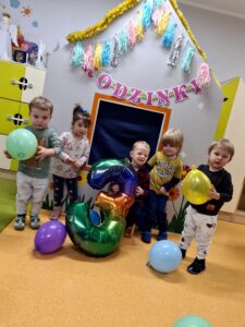 Dzieci stoją i pozują do zdjęcia z solenizantem, który siedzi na krzesełku i trzyma w dłoniach kolorowy, duży balon w kształcie cyfry trzy. W tle widać szary domek ozdobiony urodzinowymi dekoracjami.