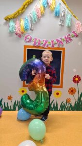 Chłopiec stoi i trzyma w dłoniach duży kolorowy balon w kształcie cyfry trzy. W tle widać szary domek z kolorowymi kwiatkami. Na domku zawieszone są urodzinowe dekoracje: różowy napis " Urodzinki" i girlanda z kolorowych pomponów.