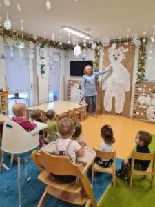 Dzieci siedzą na krzesełkach i patrzą na dużego niedźwiedzia polarnego namalowanego na szarym papierze.